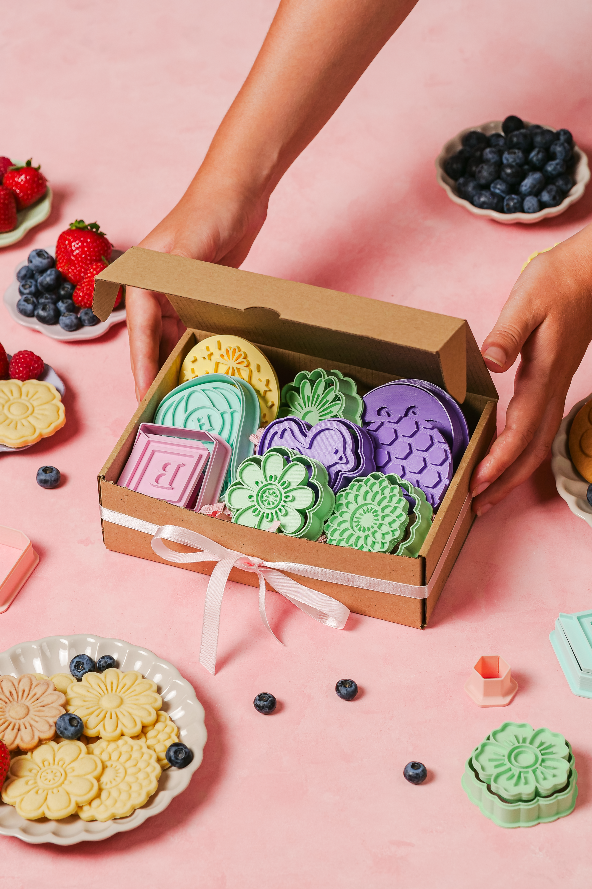 Hands holding a gift box with ribbon, filled with different formini cookie cutters and stamps in pastel colors.