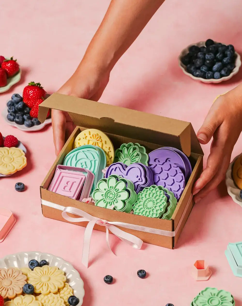 Hands holding a gift box filled with various Formini cookie cutters and stamps, there are also some prepared cookies and fruit around it
