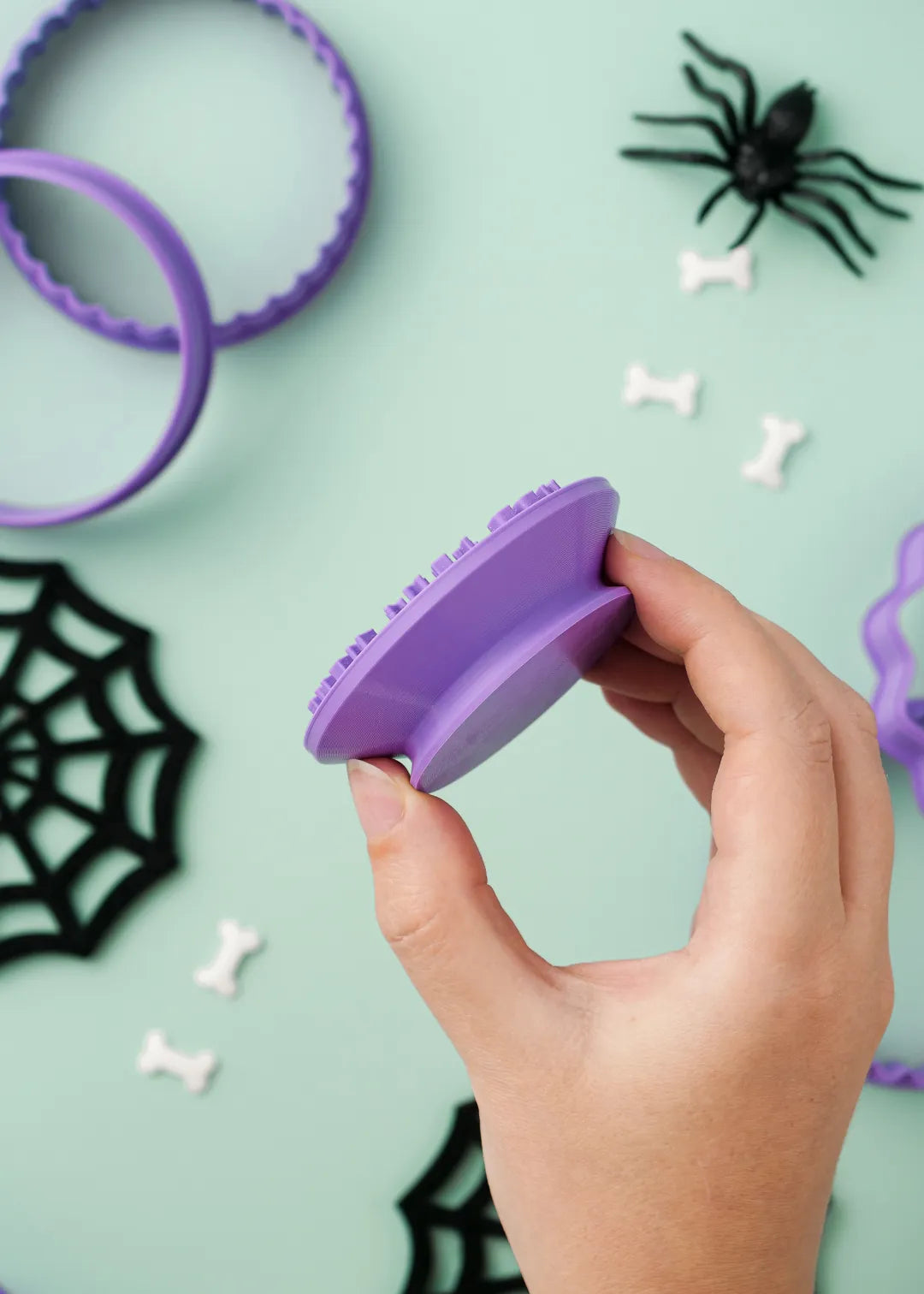 Hand holding a purple cookie stamp to show it's handle design with Halloween decorations on a light green background
