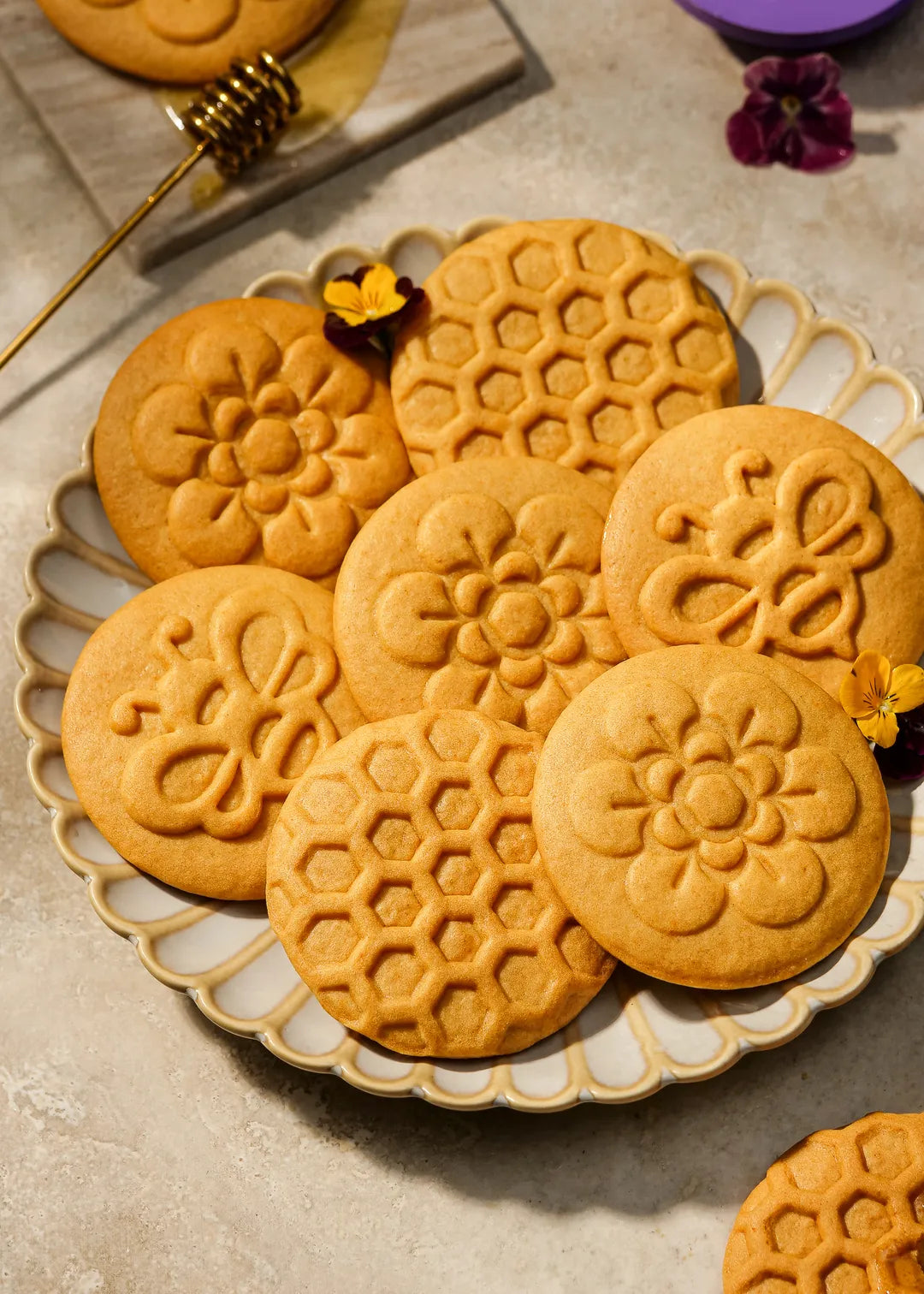 Golden-baked honey cookies with detailed embossed designs, including a honeycomb pattern, bee, and flower motifs. Displayed on a marble slab with honey drizzled around.