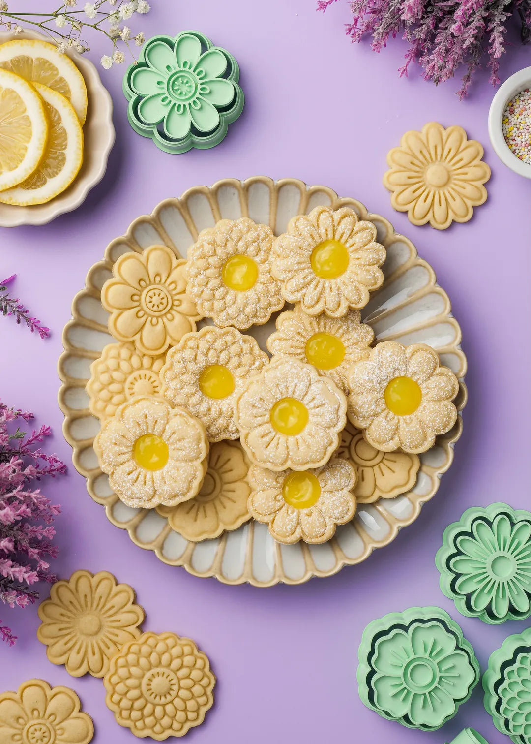 Flower-shaped linzer cookies with intricate embossed patterns, filled with vibrant yellow lemon curd, arranged on a decorative ceramic plate. Some cookies are dusted with powdered sugar. Surrounding the plate are cookie cutters, extra cookies, a small bowl of sprinkles, and a dish with fresh lemon slices. The pastel purple background is adorned with delicate lavender and white flowers.