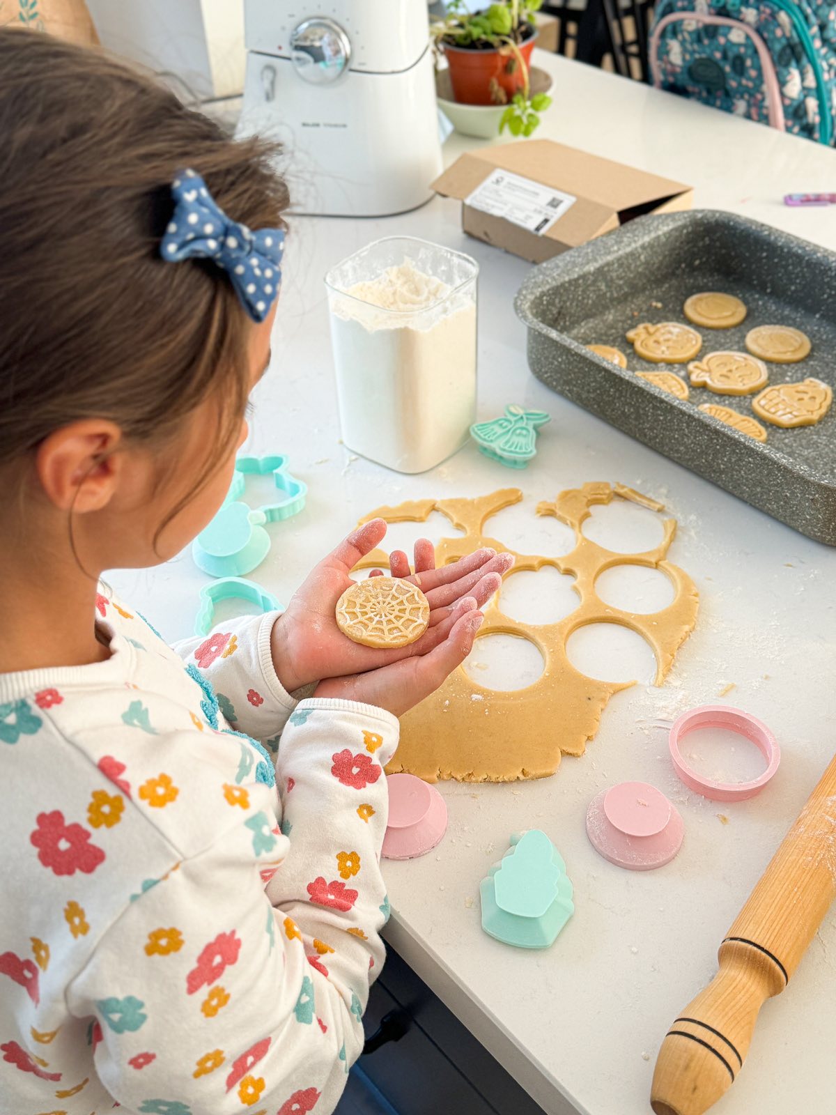 A girl holding a stamped cookie in her hands as she's preparing cookies. There is a rolled out cookie dough, a baking sheet filled with more stamped cookies, and various Formini cookie cutters and stamps on the countertop. The signature ergonomic grip handles of the stamps can be seen.