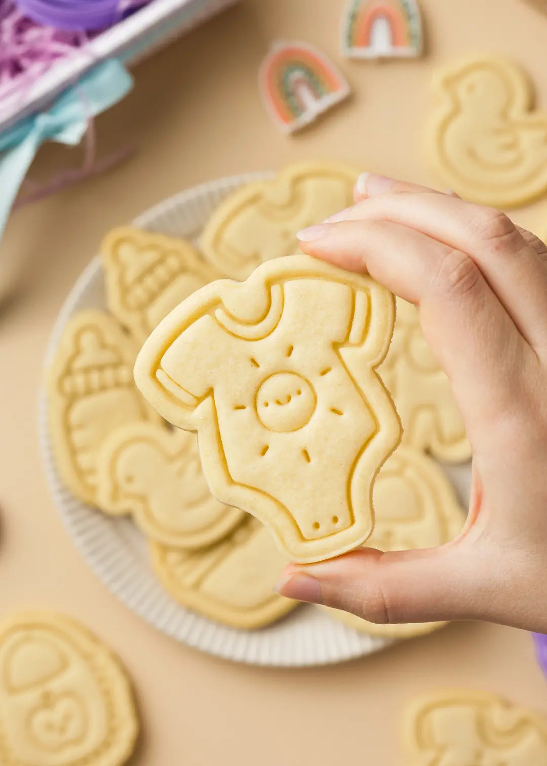 A close-up of a hand holding a baby onesie-shaped sugar cookie with a sun and smiley face embossed design