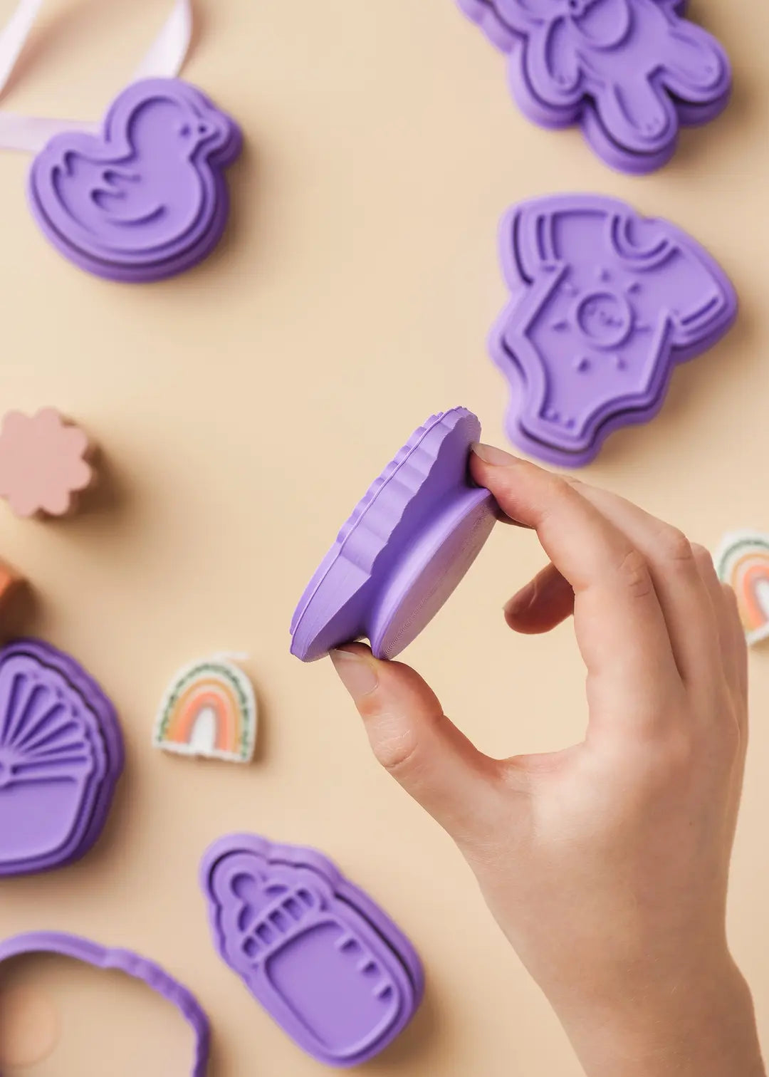 Close-up of a hand holding a purple baby-themed cookie cutter with a special made comfortable grip. High-quality 3D-printed cookie stamp for baby shower treats and baby parties. Perfect gift for bakers.