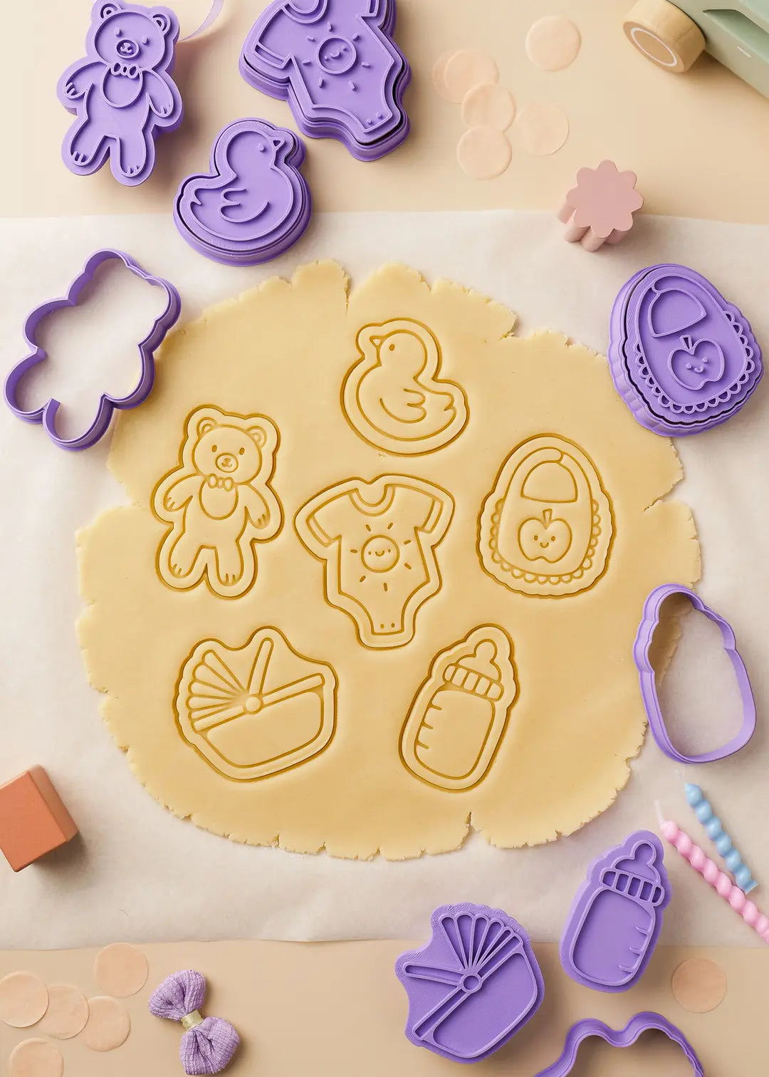 Baby-themed cookie cutters pressing adorable shapes into rolled cookie dough. Includes teddy bear, baby bottle, onesie, duck, bib, and bassinet designs for baby shower baking.