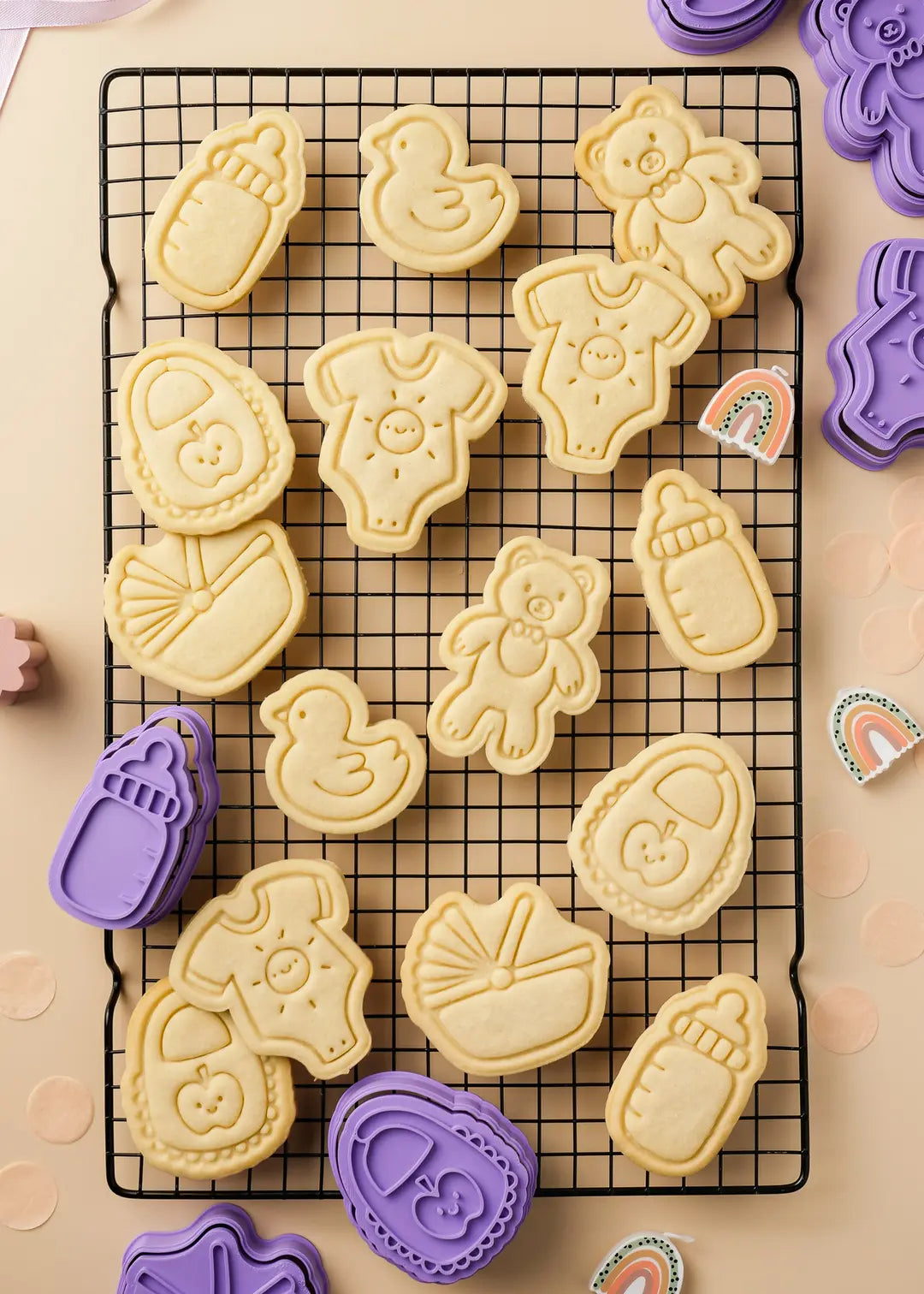 A cooling rack with baked baby shower cookies, decorated with adorable embossed designs, alongside purple cookie cutters.