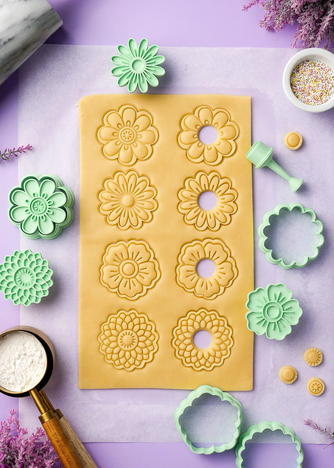 Floral-shaped cookie dough cutouts arranged on parchment paper, ready for baking. Mint Green flower cookie cutters are scattered around, showcasing intricate embossed patterns. A soft pastel background enhances the delicate baking scene.