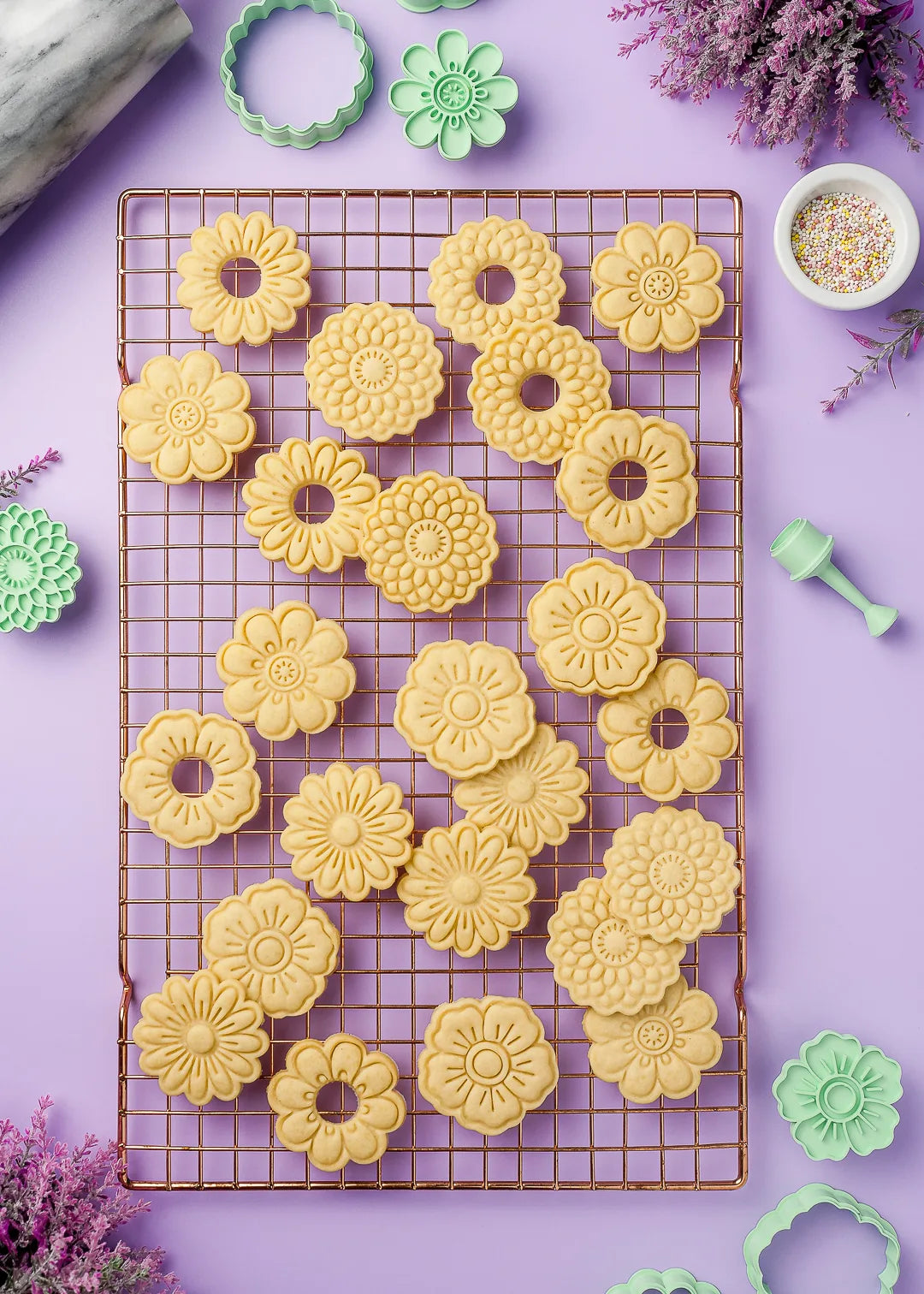 Floral-patterned cookies shaped using plastic cookie cutters, arranged on a copper cooling rack. Surrounding them are various cookie cutters, a marble rolling pin, and a small bowl of colorful sprinkles. 
