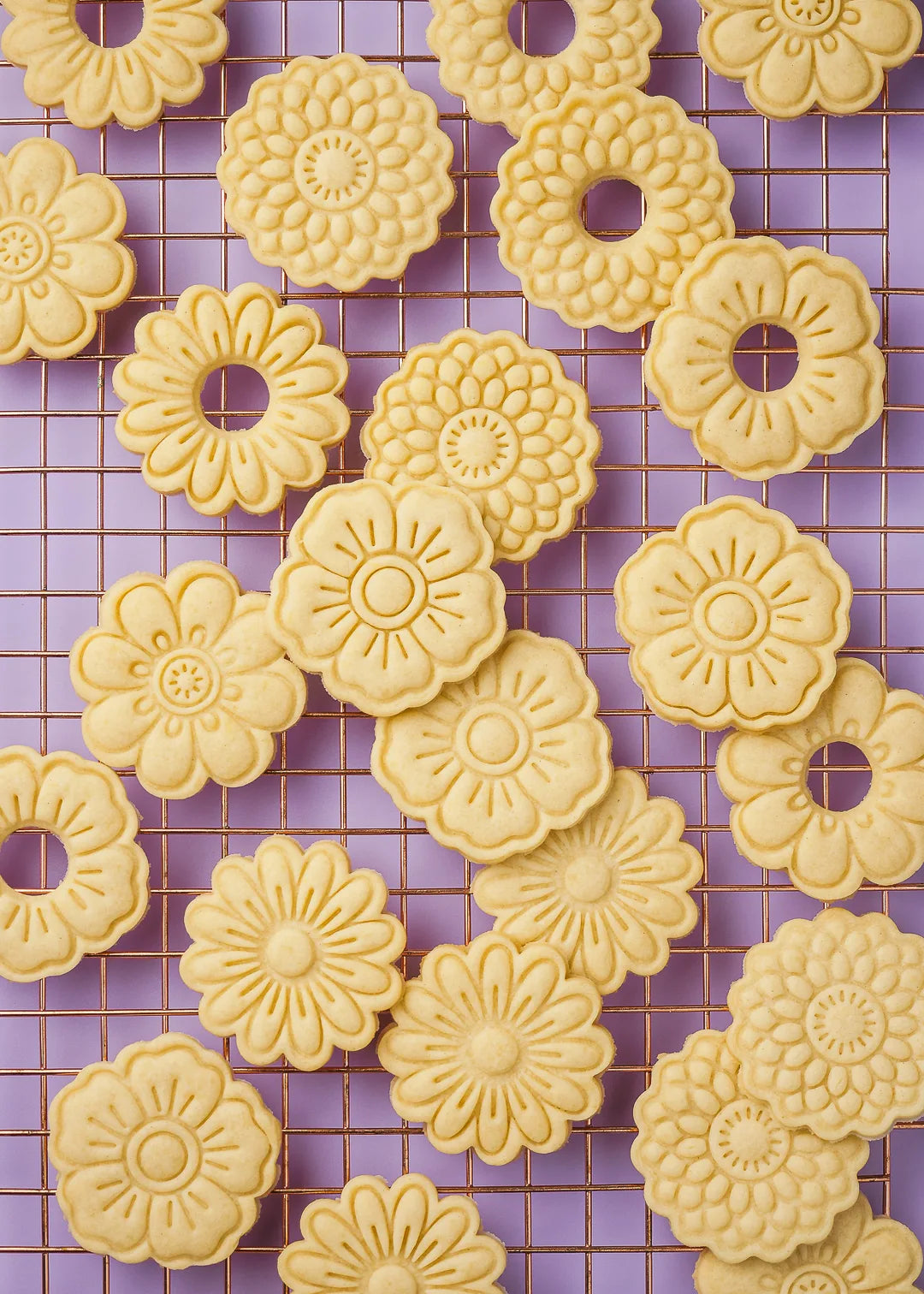 Floral-patterned cookies shaped using plastic cookie cutters, arranged on a copper cooling rack. 