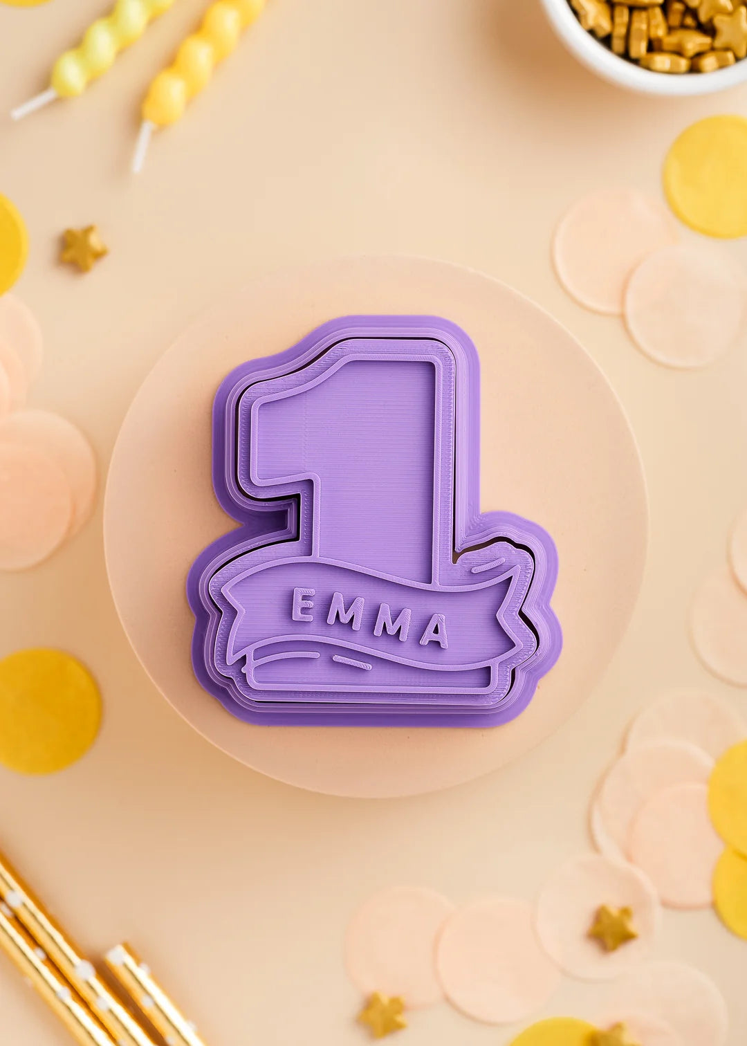 Purple number one cookie stamp and cutter with custom name 'Emma', designed for a first birthday celebration. Surrounded by pastel confetti and party decorations.