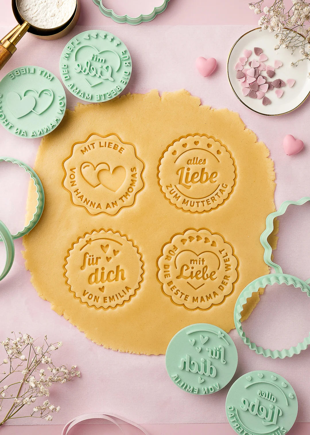 Stamped cookie dough with different German messages such as “für dich – von Emilia,” “mit Liebe – von Hanna an Thomas,” “alles Liebe zum Muttertag,” and “für die beste Mama der Welt.” Surrounding the dough are mint green cookie stamps and cutters, all arranged on a pink baking surface with decorative flowers and heart sprinkles. A perfect example of how the stamps leave clear, beautiful impressions in the dough.