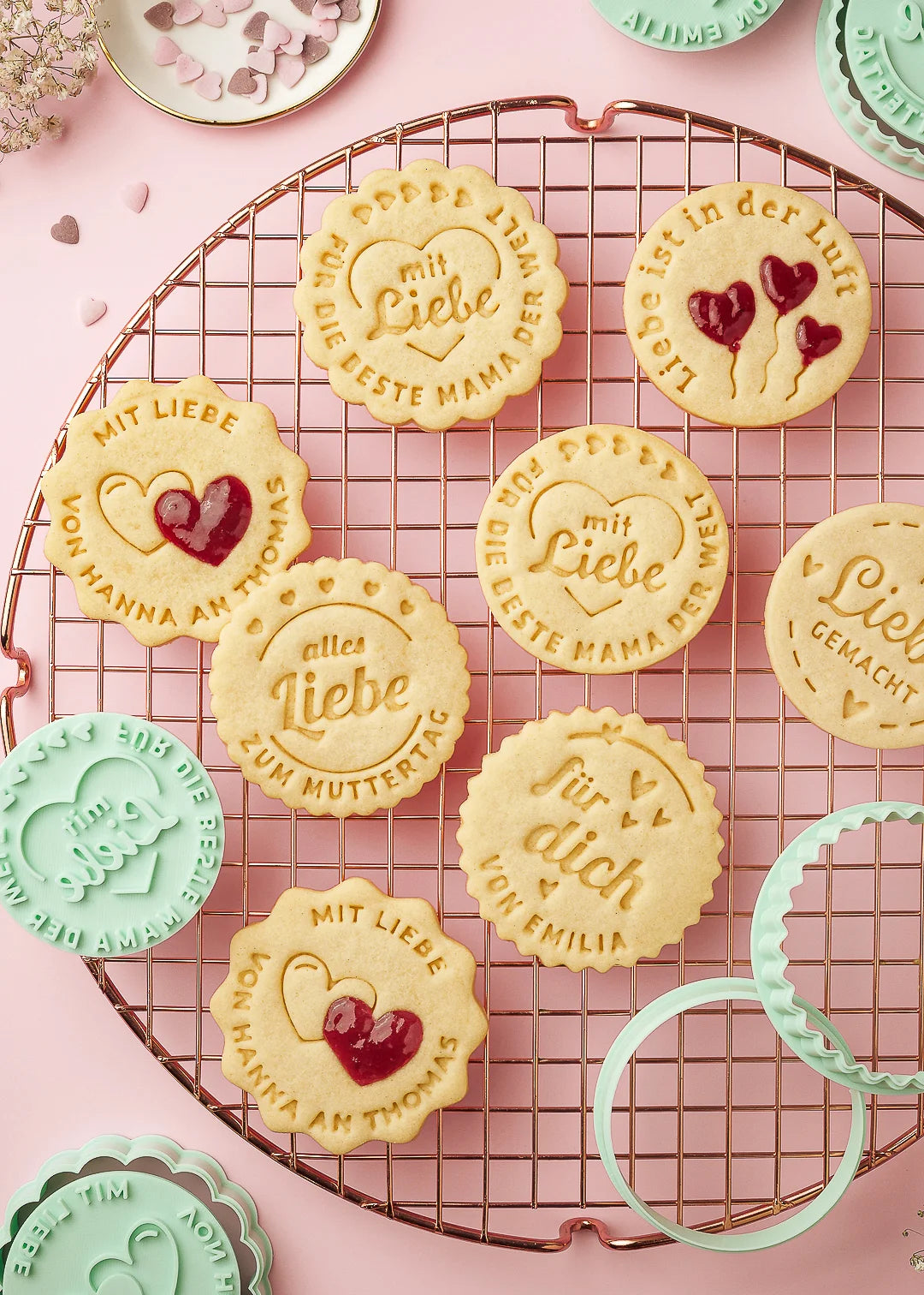 A collection of beautifully stamped cookies with German messages like “mit Liebe,” “für dich – von Emilia,” and “Liebe ist in der Luft,” displayed on a cooling rack. Some cookies feature red jam-filled heart designs. Surrounding the cookies are mint green cookie stamps and matching cutters, all made with precision. A perfect gift idea and ideal for personalized baking using Formini cookie stamps.
