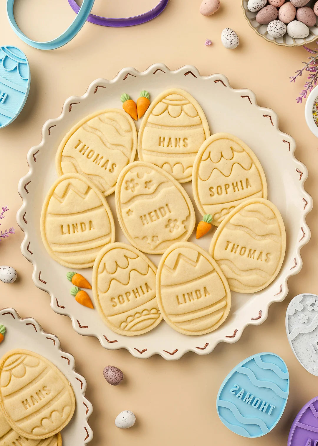 Baked Easter egg cookies personalized with different names and arranged on a plate, with cookie cutters and stamps around it