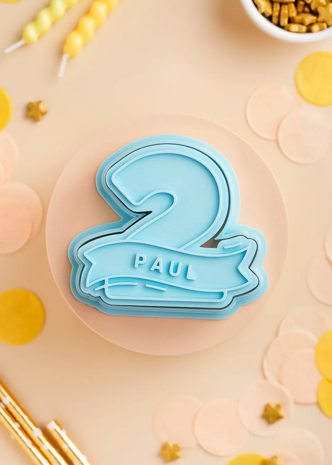 Pastel Blue number two cookie stamp with cutter with custom name 'Paul', perfect for a kids birthday party. Surrounded by pastel confetti and party-themed decorations.
