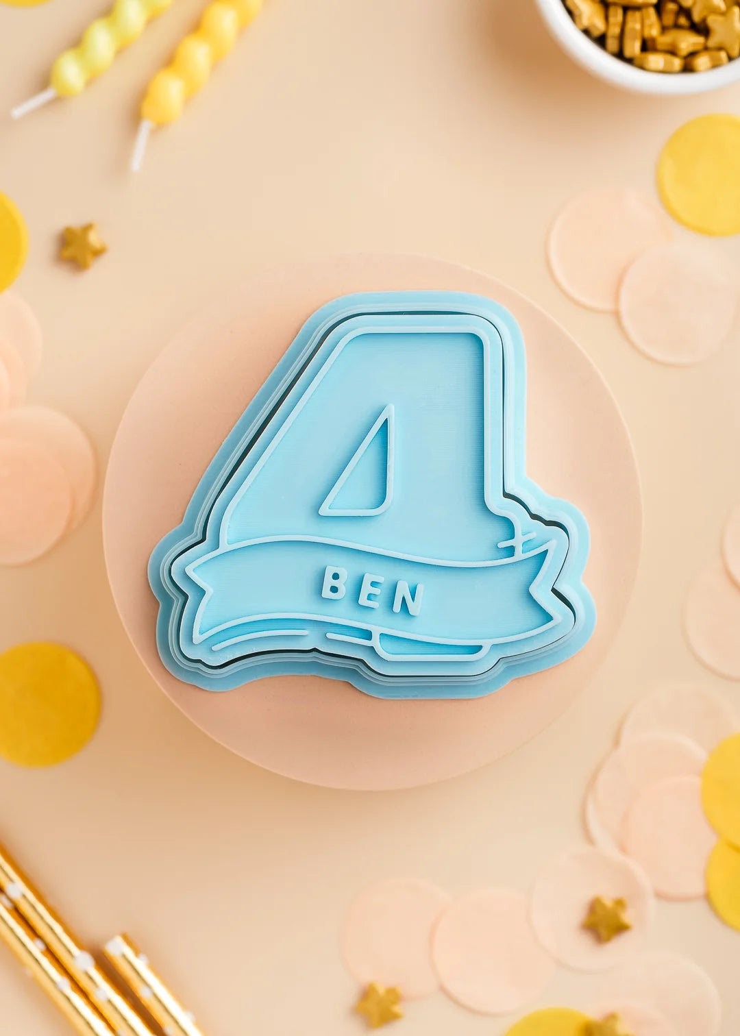 Pastel Blue number four cookie stamp with cutter with custom name 'Ben', perfect for a kids birthday party. Surrounded by pastel confetti and party-themed decorations.
