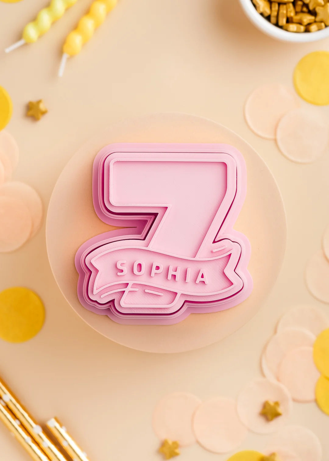 Pastel Pink number seven cookie stamp with cutter with custom name 'Sophia', perfect for a kids birthday party. Surrounded by pastel confetti and party-themed decorations.