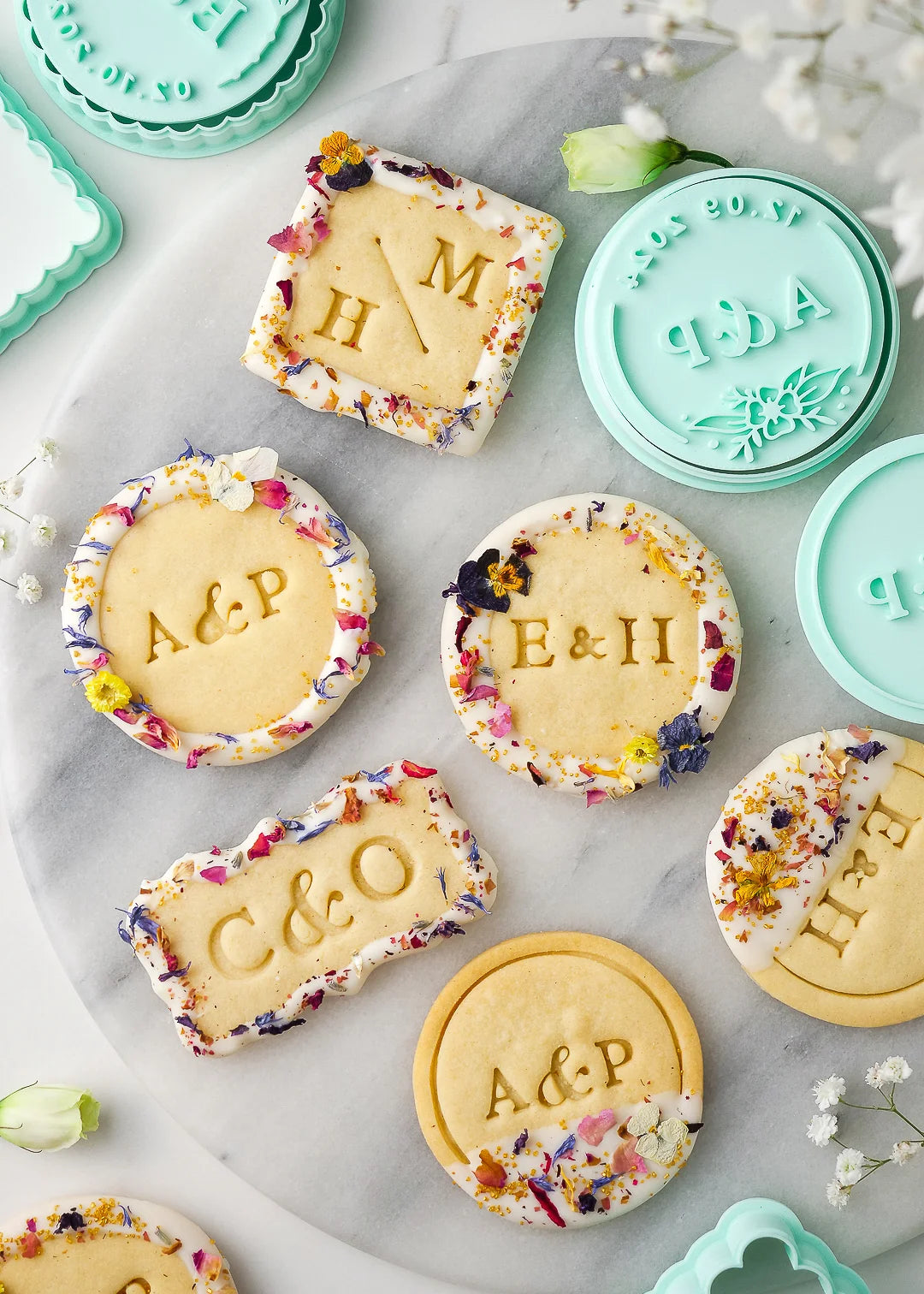 Wedding-themed personalized cookies displayed on a marble platter, showcasing various custom cookie stamp designs including round, square, and rectangular shapes. Each cookie features options like initials, wedding date, couple’s names, or thank you messages, made with Formini’s customizable cookie cutters and stamps. The cookies are decorated with white chocolate and dried flowers.
