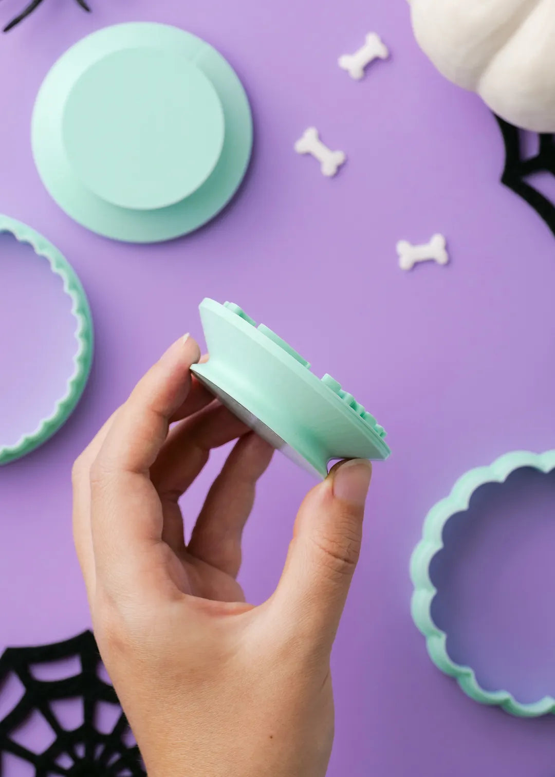 Hand holding a green cookie stamp with handle against a purple background with Halloween-themed decorations.