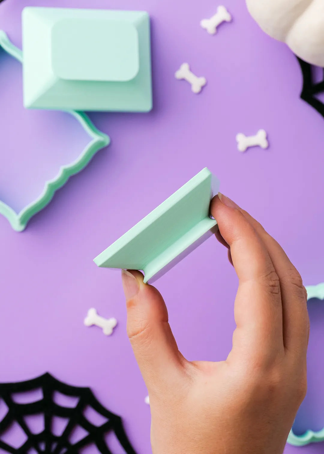 Hand holding a green cookie stamo against a purple background with Halloween decorations.