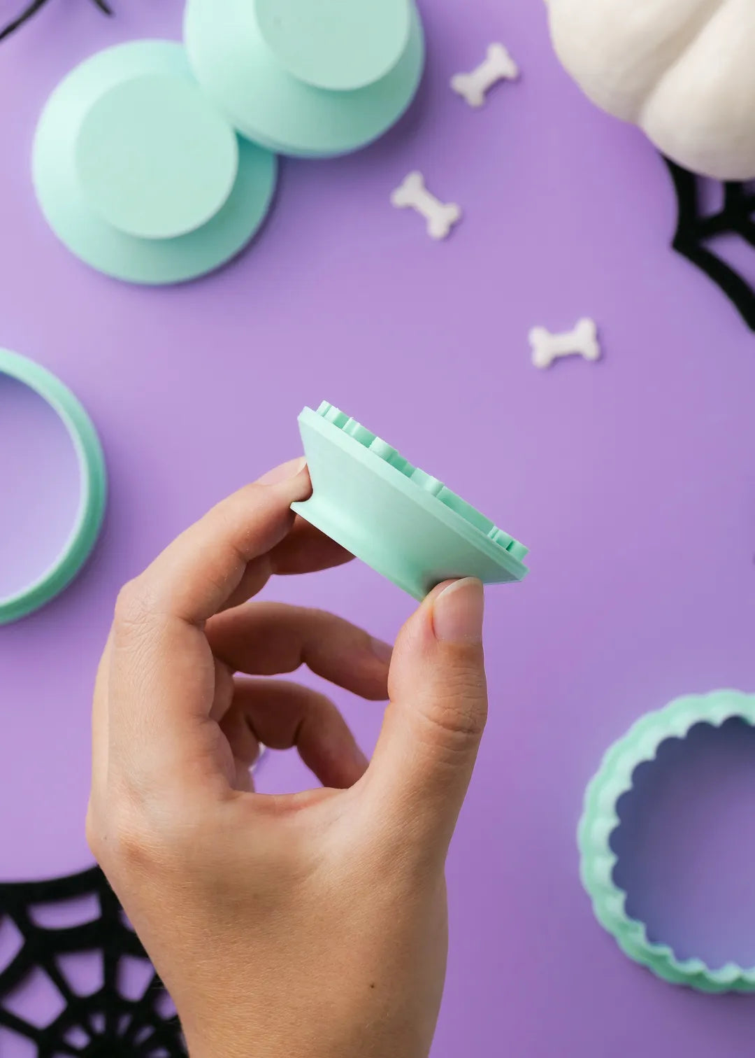 Hand holding a green cookie stamp against a purple background with cookie cutters and bone-shaped cookies.