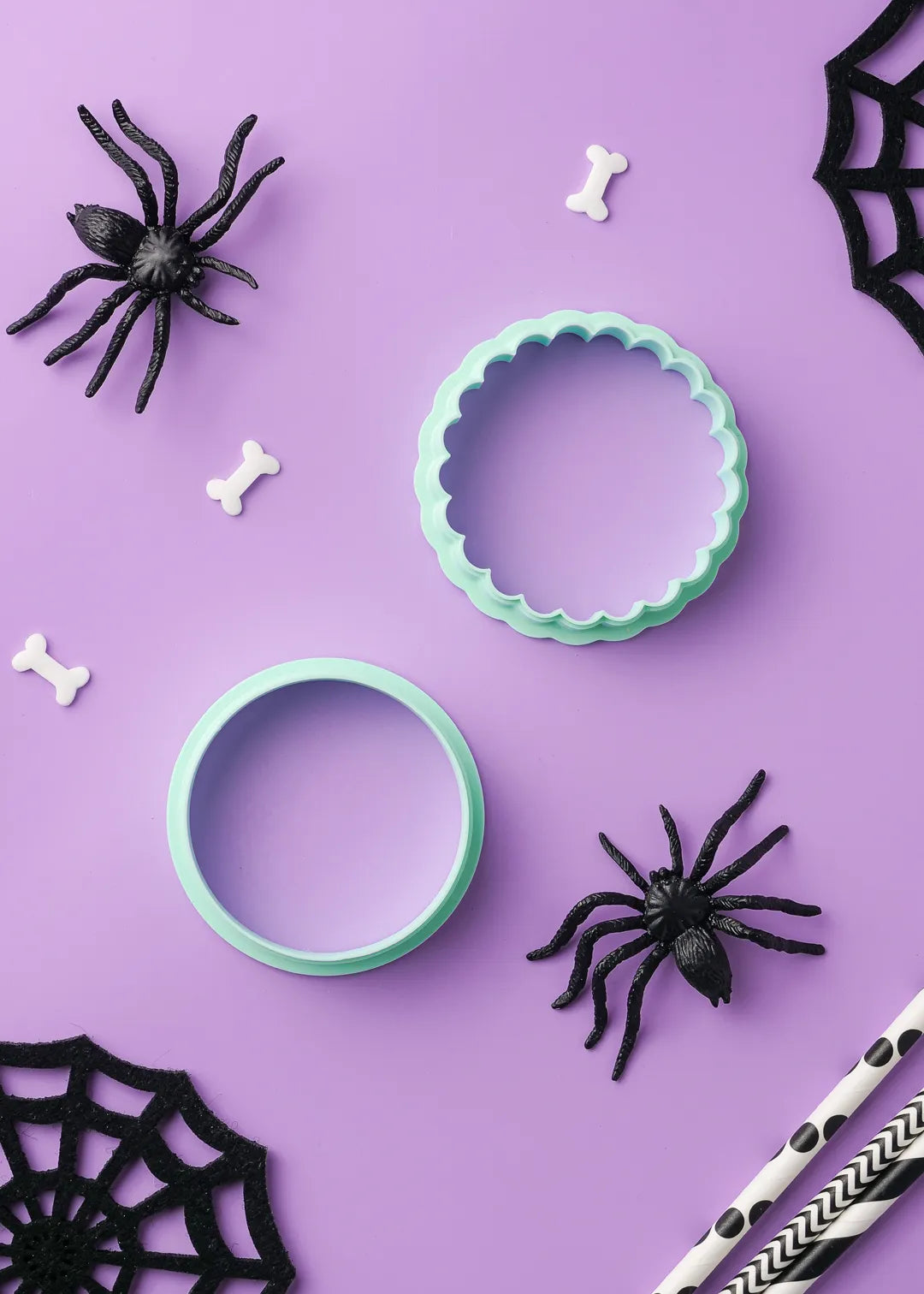 Two green cookie cutters on a purple background with Halloween decorations including black spiders and spider webs. Cookie cutters are - plain circle and scalloped circle