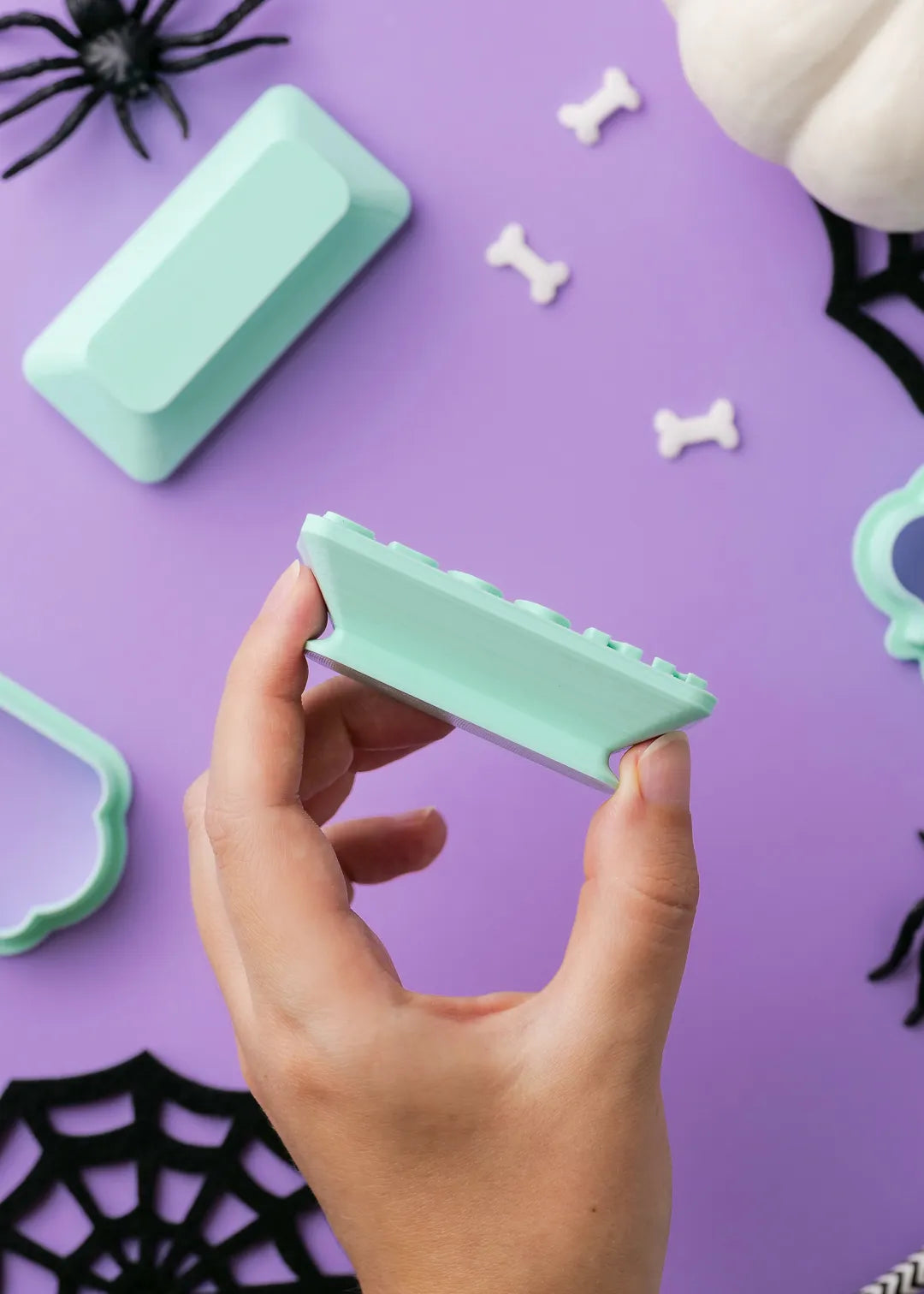 Hand holding a green cookie stamp to show it's ergonomic handle design against a purple background with Halloween decorations.