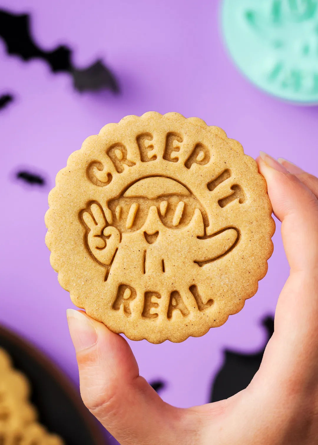 Cookie with 'Creep It Real' text ang cool ghost design held by a hand against a purple background with bats.