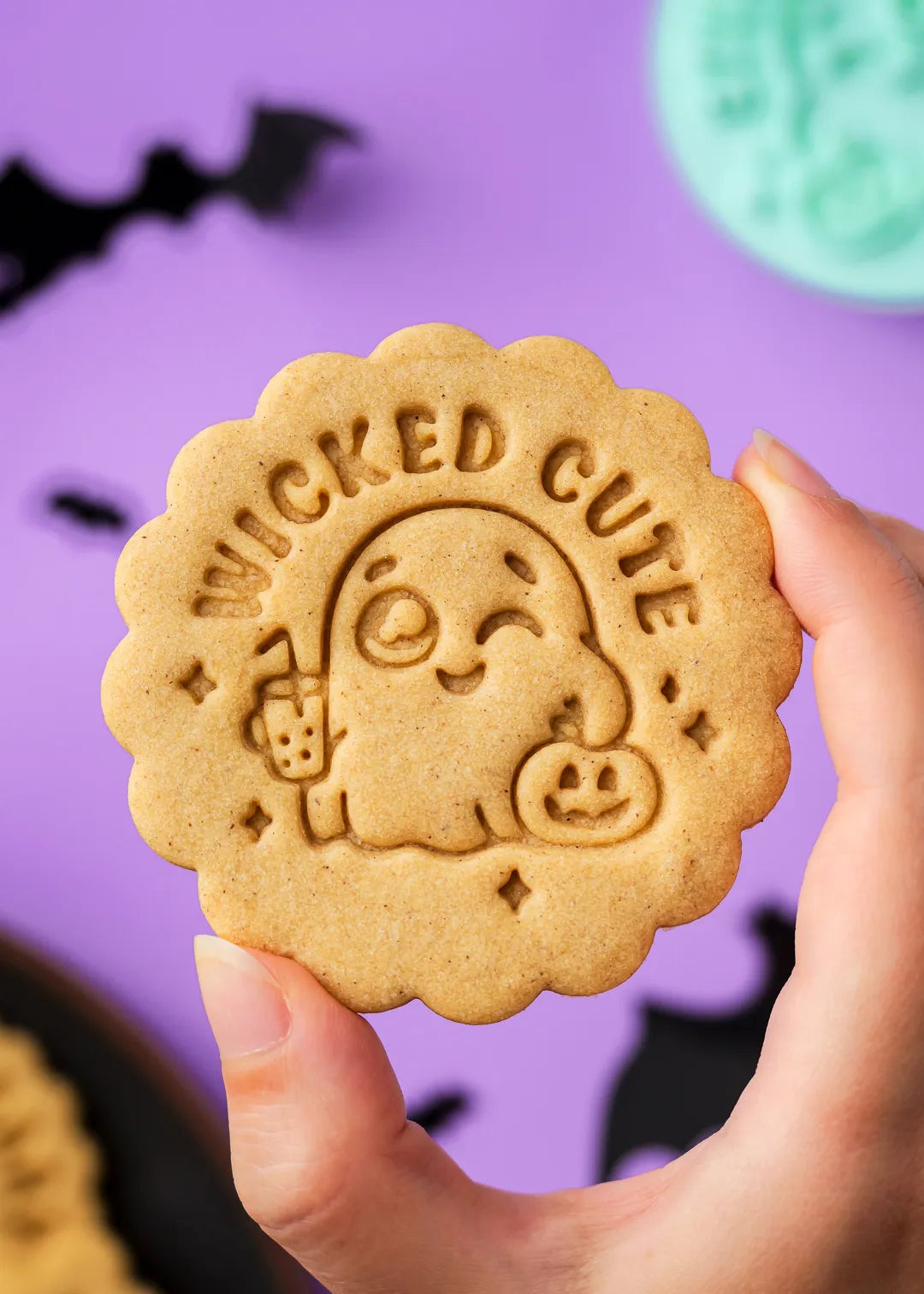 Hand holding a cookie with 'Wicked Cute' text and ghost holding a cup and pumpkin design against a purple background