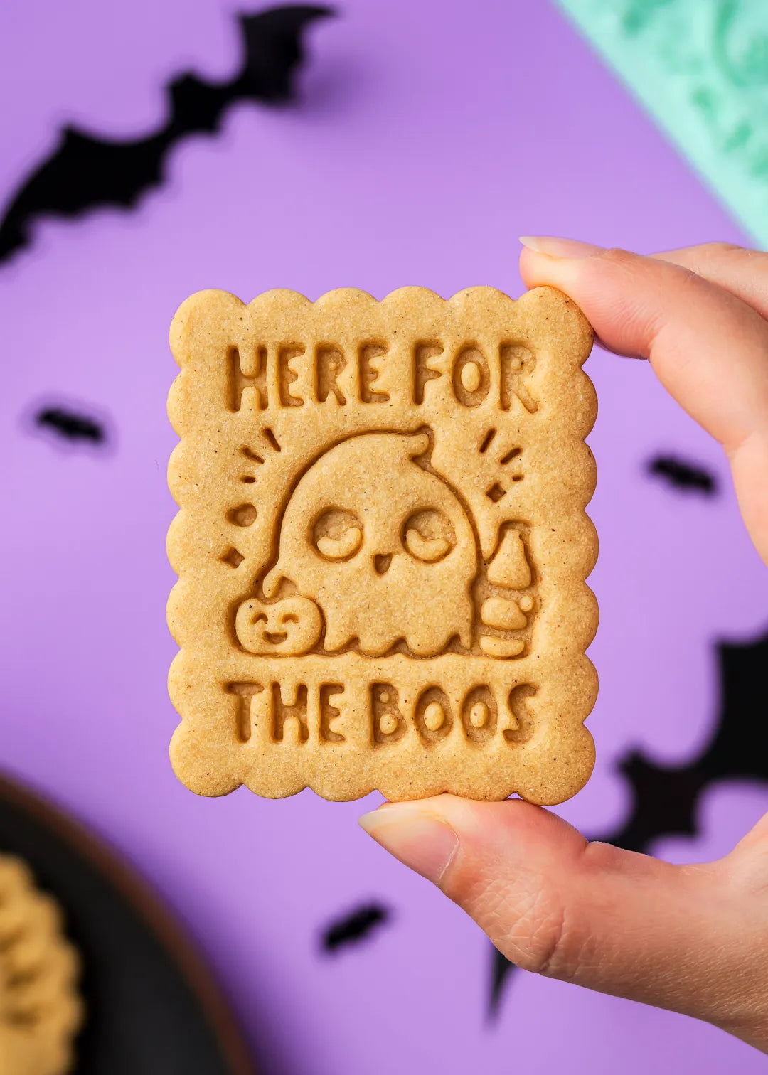 Halloween cookie with 'Here for the Boos' text held by a hand against a purple background with bats.