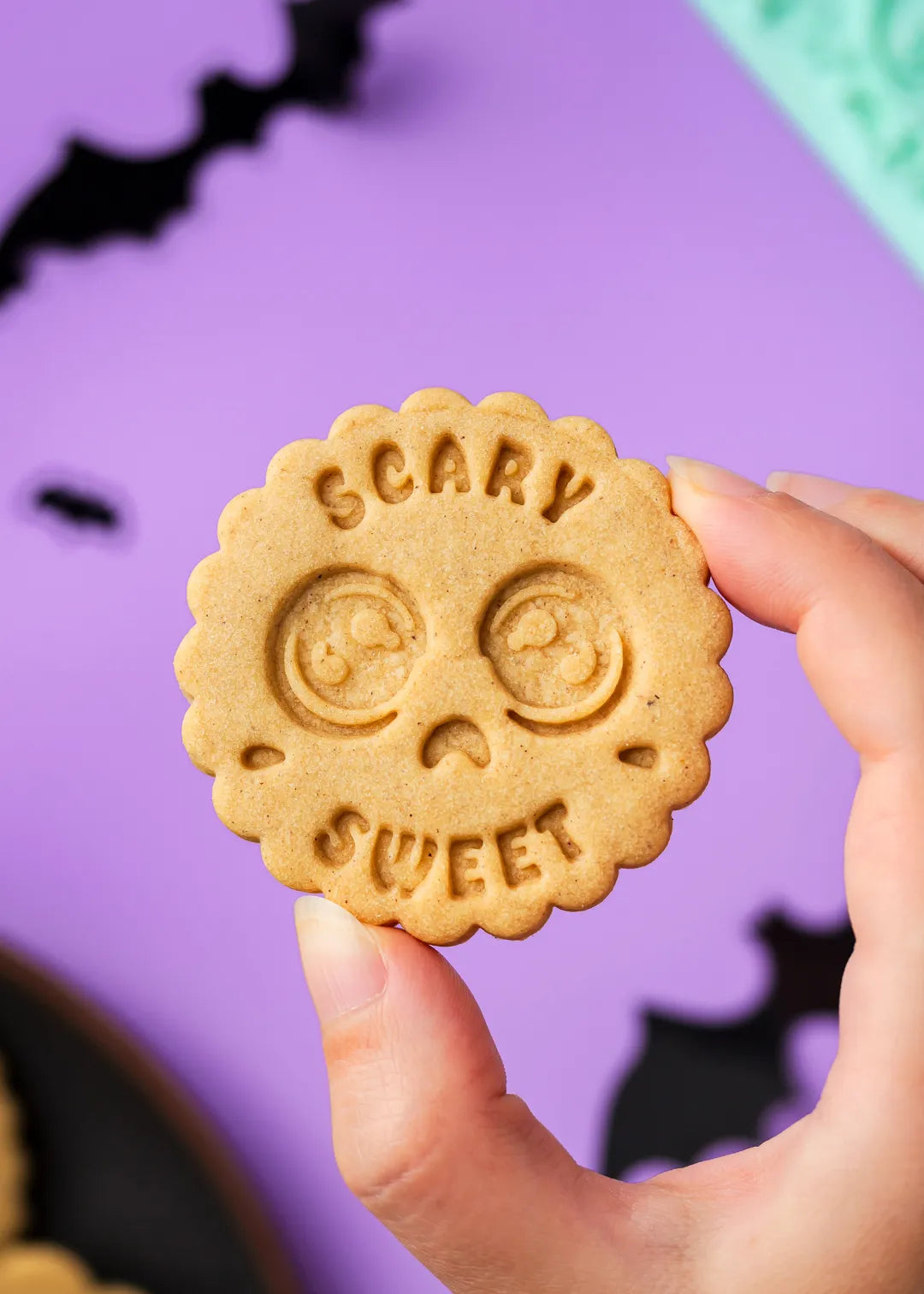 Hand holding a cookie with 'Scary Sweet' text against a purple background with black bats.