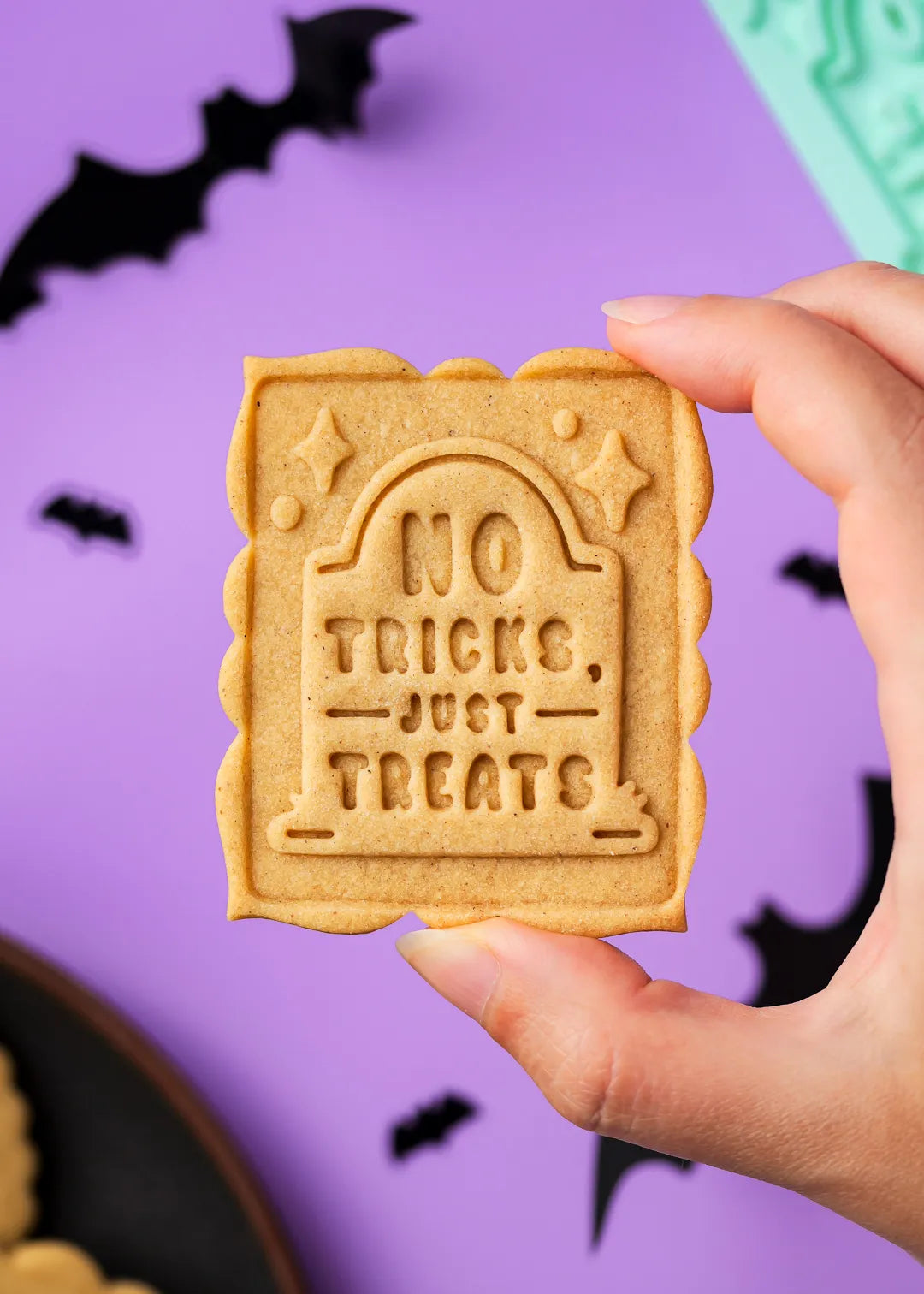 Hand holding a Halloween-themed cookie with 'No Tricks, Just Treats' text against a purple background with bats.