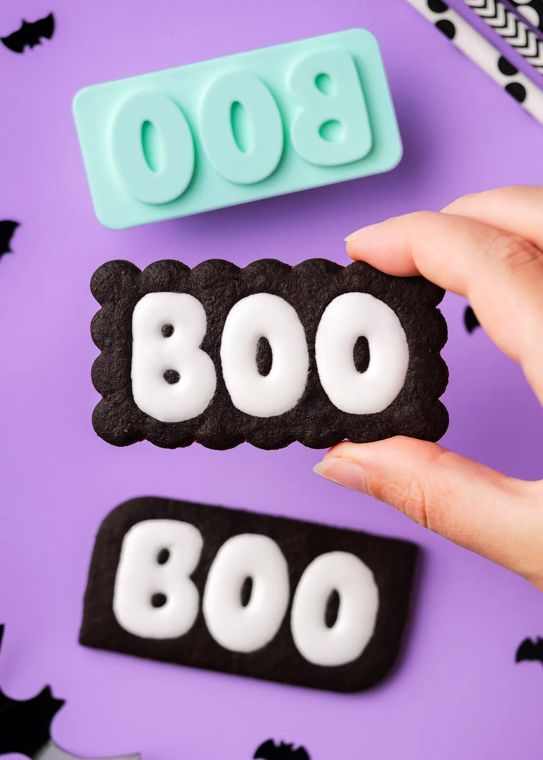 Hand holding a 'BOO' chocolate cookie with a cookie stamp in the background on a purple surface. The boo text is filled with vanilla icing.