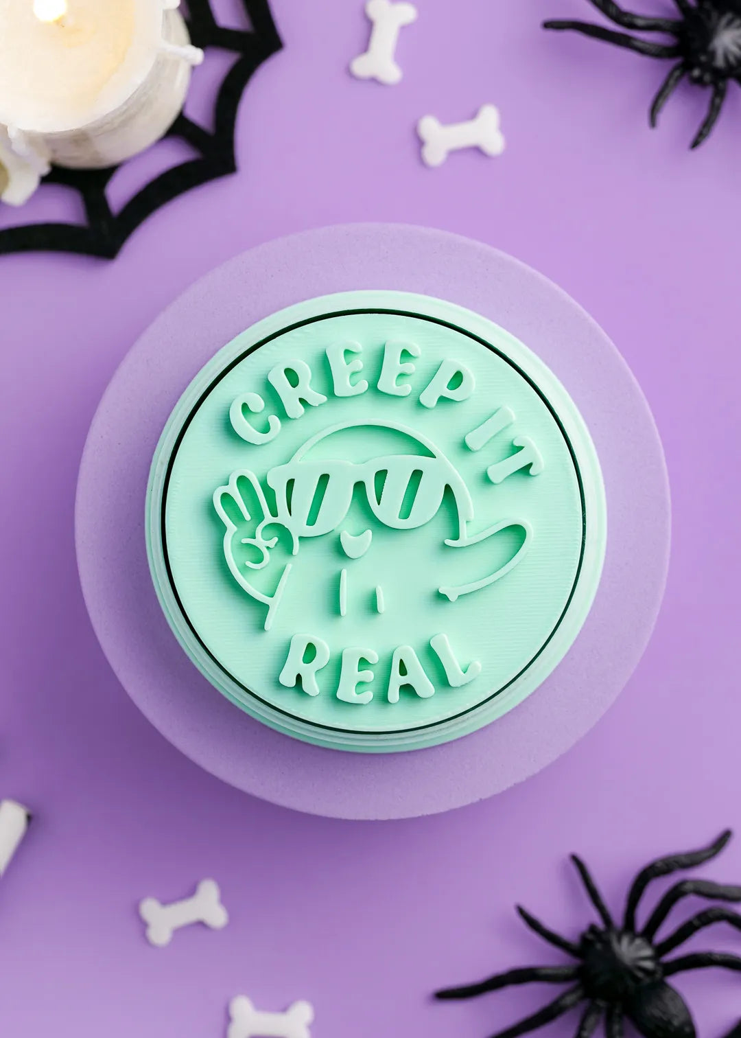 Mint green stamp with 'Creepy It's Real' text and cute ghost design on a purple background with Halloween decorations.