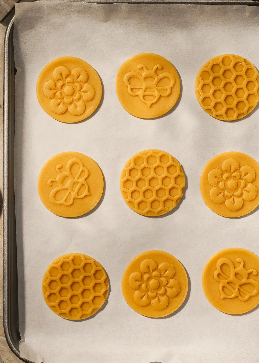 Raw honey cookie dough with bee, flower, and honeycomb patterns, perfectly shaped before baking. The embossed cookie dough is neatly arranged on a baking sheet.