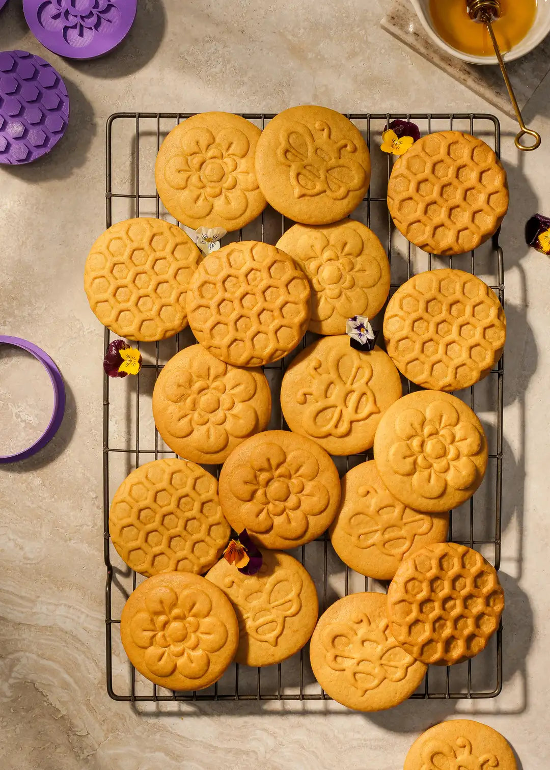 Golden-baked honey cookies with detailed embossed designs, including a honeycomb pattern, bee, and flower motifs. Displayed on a marble slab with honey drizzled around.