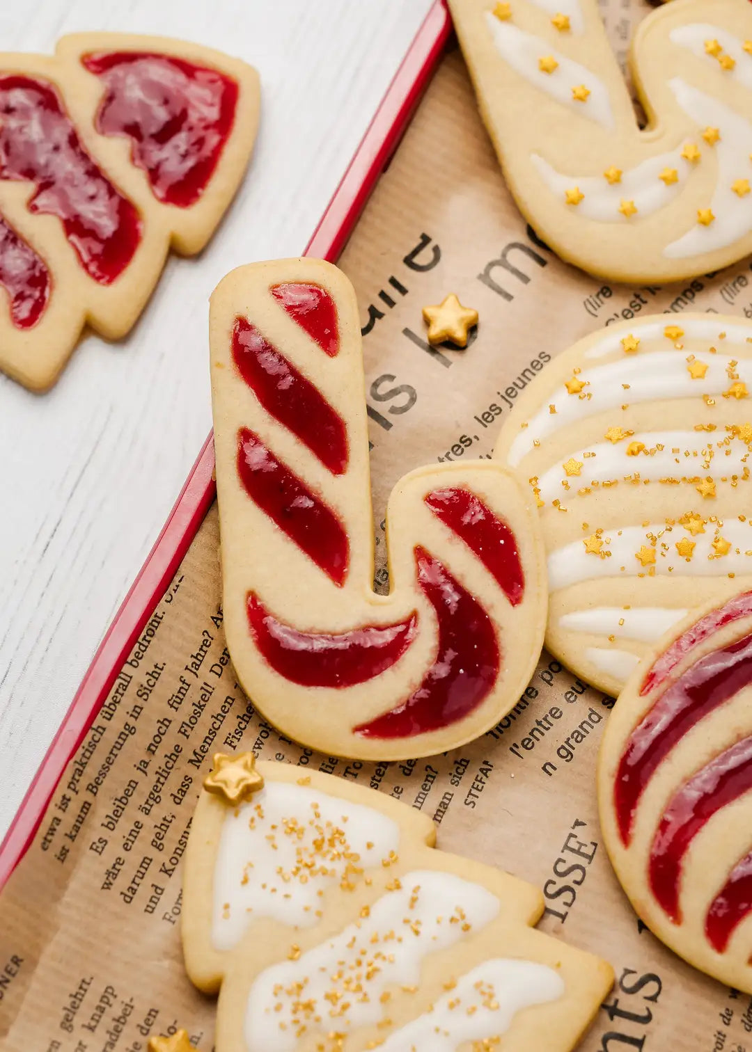 Close-up photo of a Christmas candy cane cookie made with the cookie cutter and stamp from the set. The cookie is finished jam and the other cookies around are finished with simple icing made from powdered sugar and milk, and some golden star sprinkles on top.